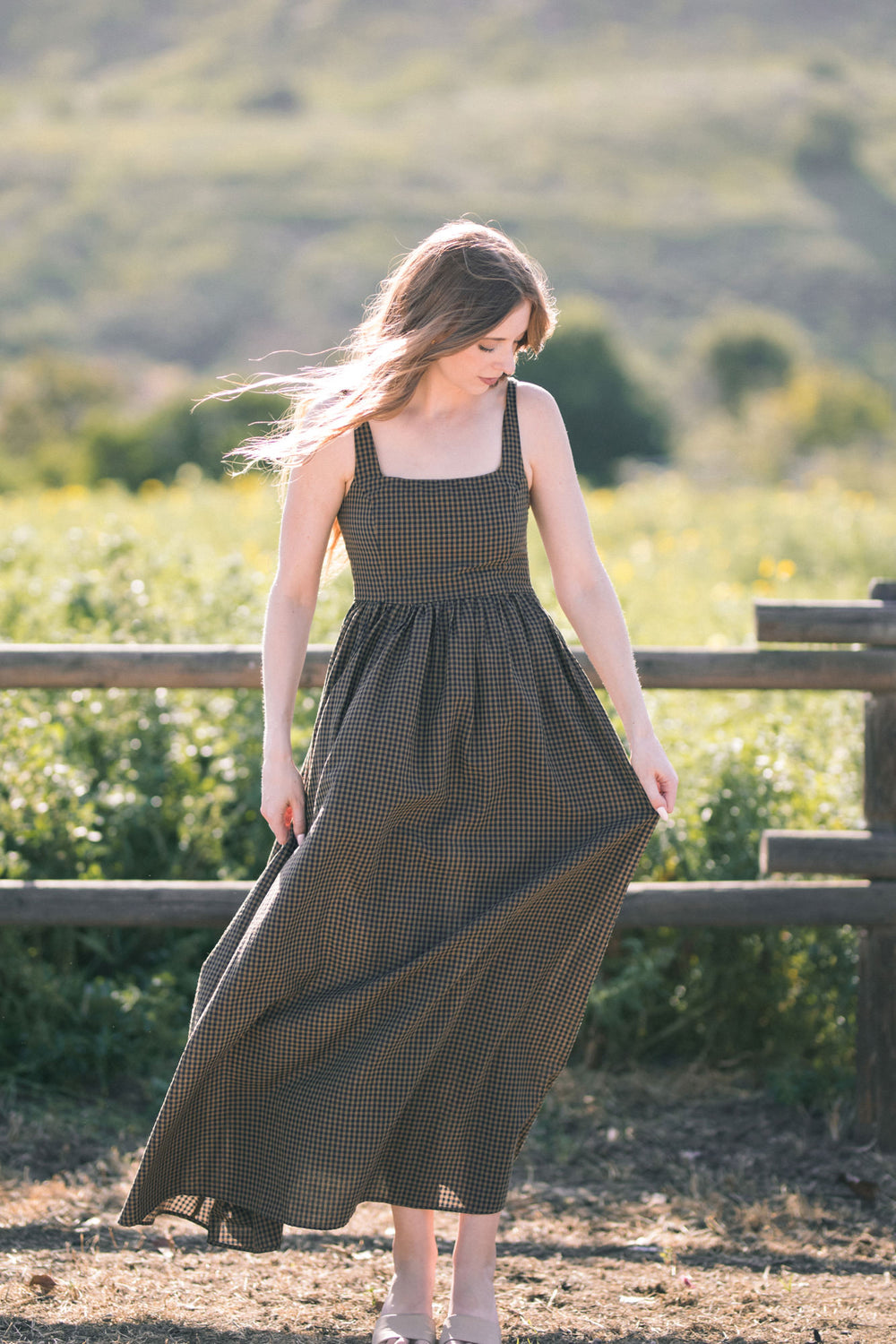 A young woman in a long, flowing black-and-white gingham maxi dress stands in a field, with lush greenery and a blurred background.