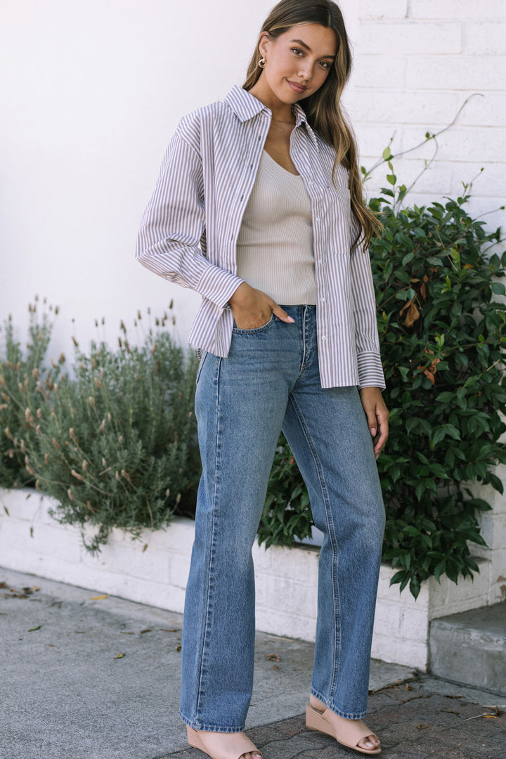 A woman wearing a striped button-up shirt with a neutral camisole and high-waisted blue jeans, standing in front of greenery.