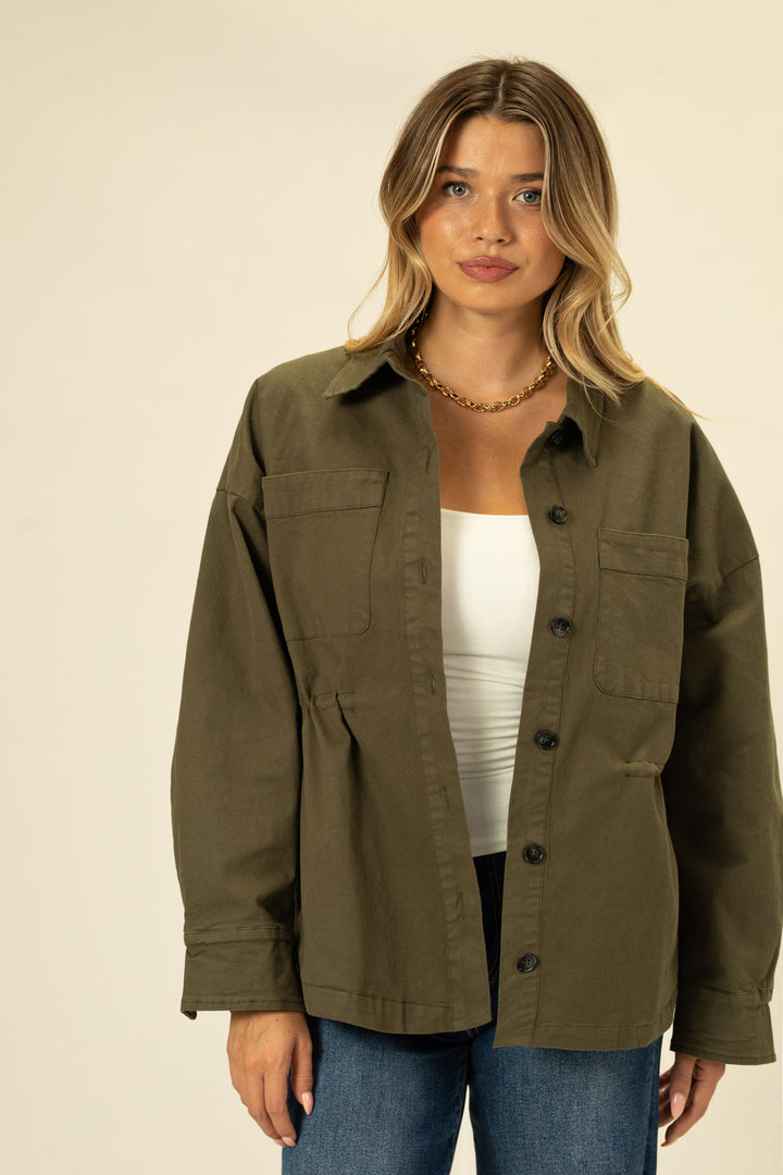 A woman in an olive green, button-up jacket with cinched waist and patch pockets, showcasing the product's style and features.