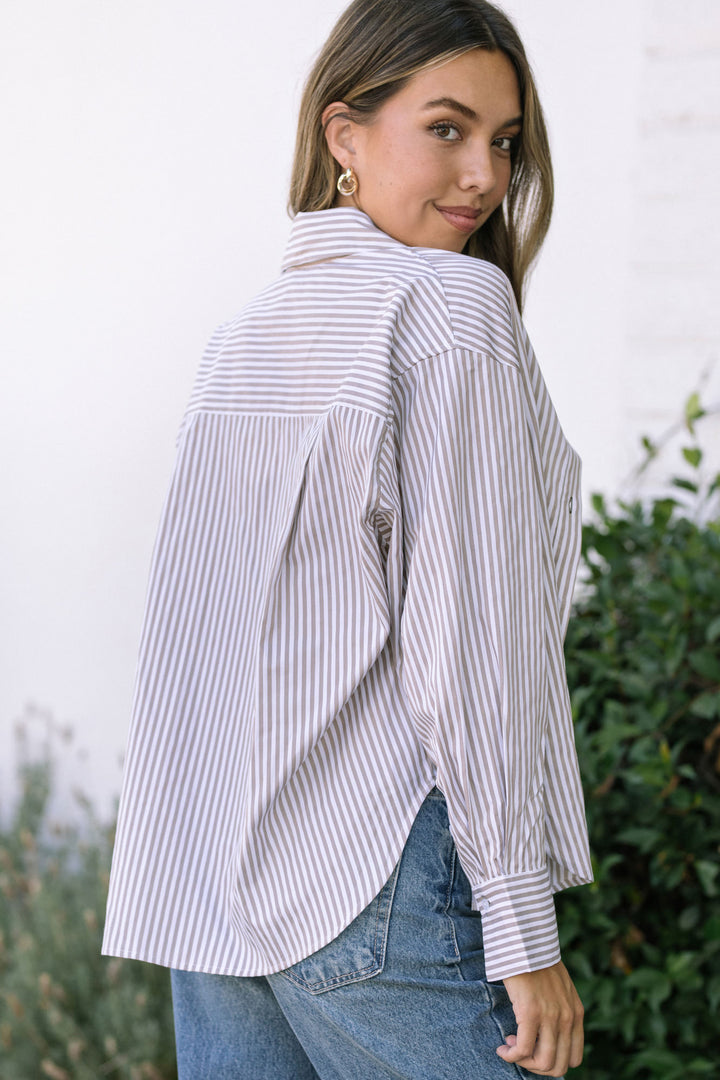 A young woman wearing a striped button-up shirt with a relaxed, casual style and flowing sleeves, posing against a natural, greenery-filled backdrop.