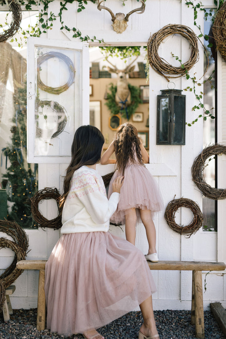 Two young women wearing pink dresses and sweaters, standing in front of a rustic white wall decorated with hanging wreaths and a deer skull.