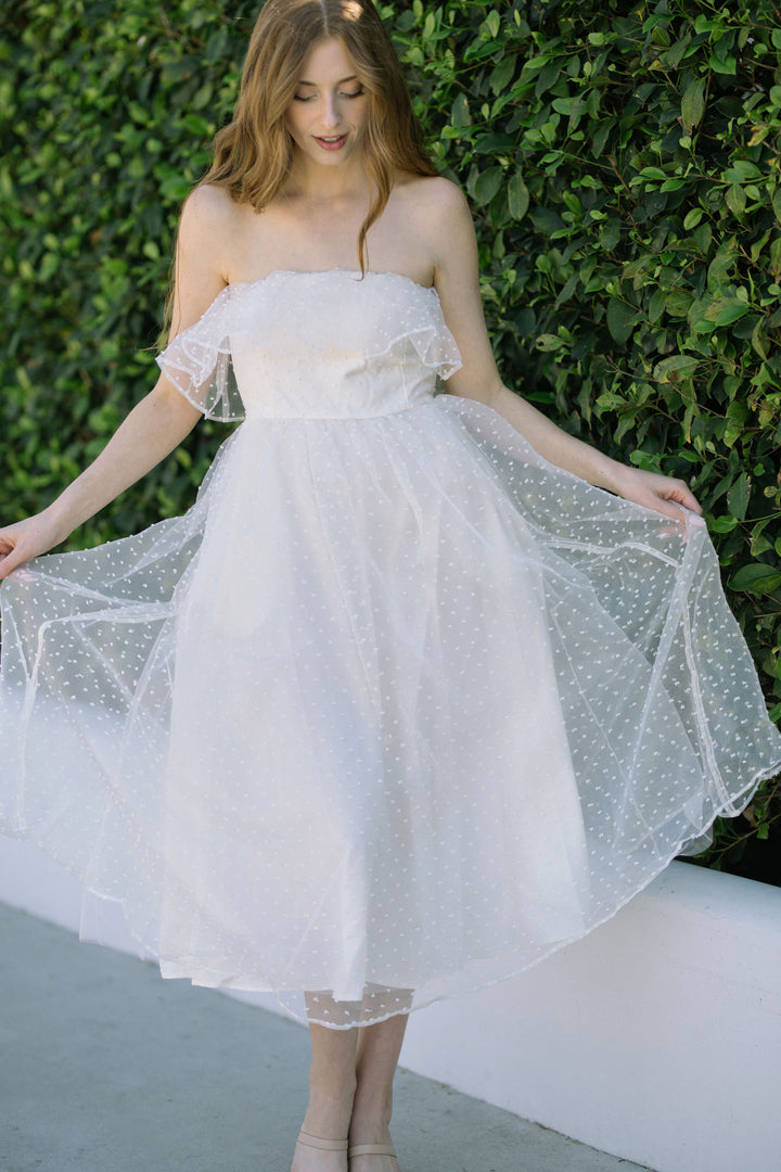 A strapless white tulle midi dress with a ruffle off-the-shoulder neckline and a full, flared skirt decorated with delicate polka dot patterns.