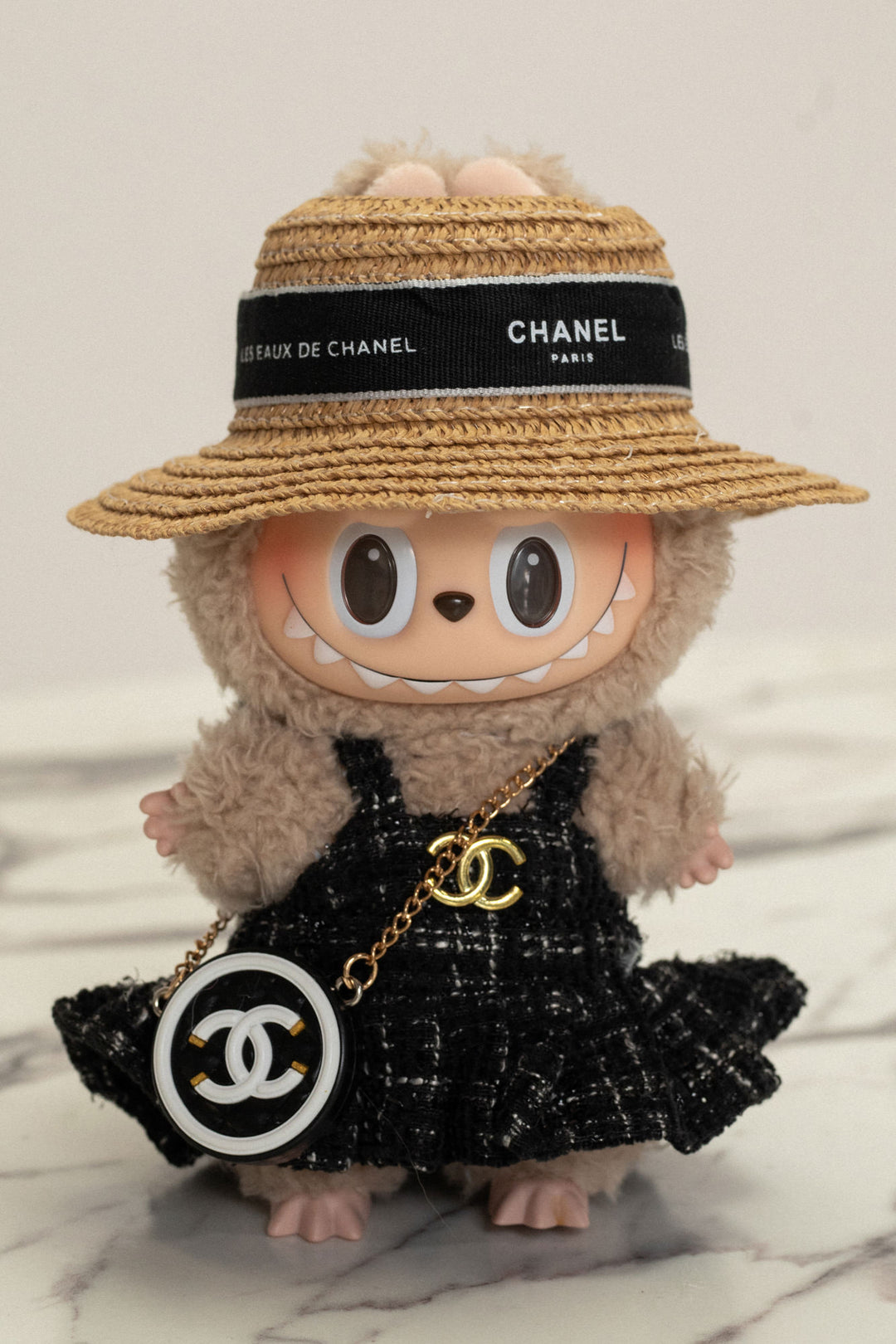 Fuzzy gray plush Labubu doll wearing a black Chanel-inspired tweed dress and straw sun hat with designer branding.