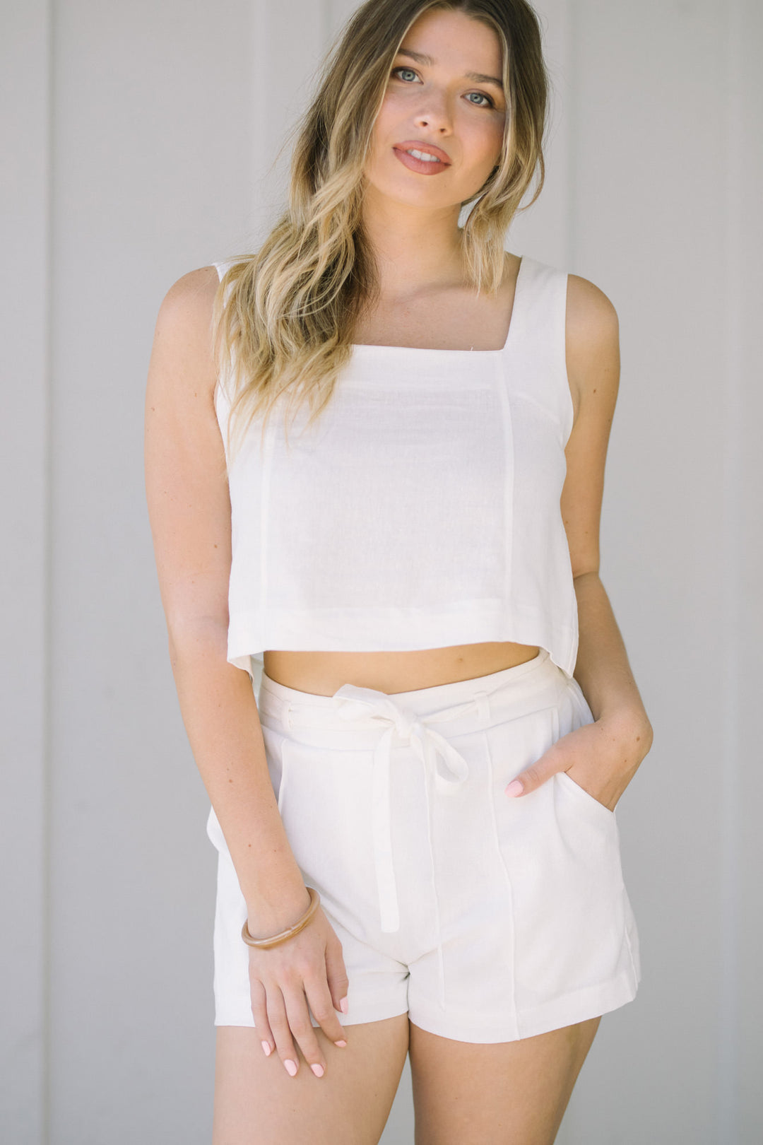 A white crop top with a square neckline and high-waisted, off-white shorts with a waist tie detail.