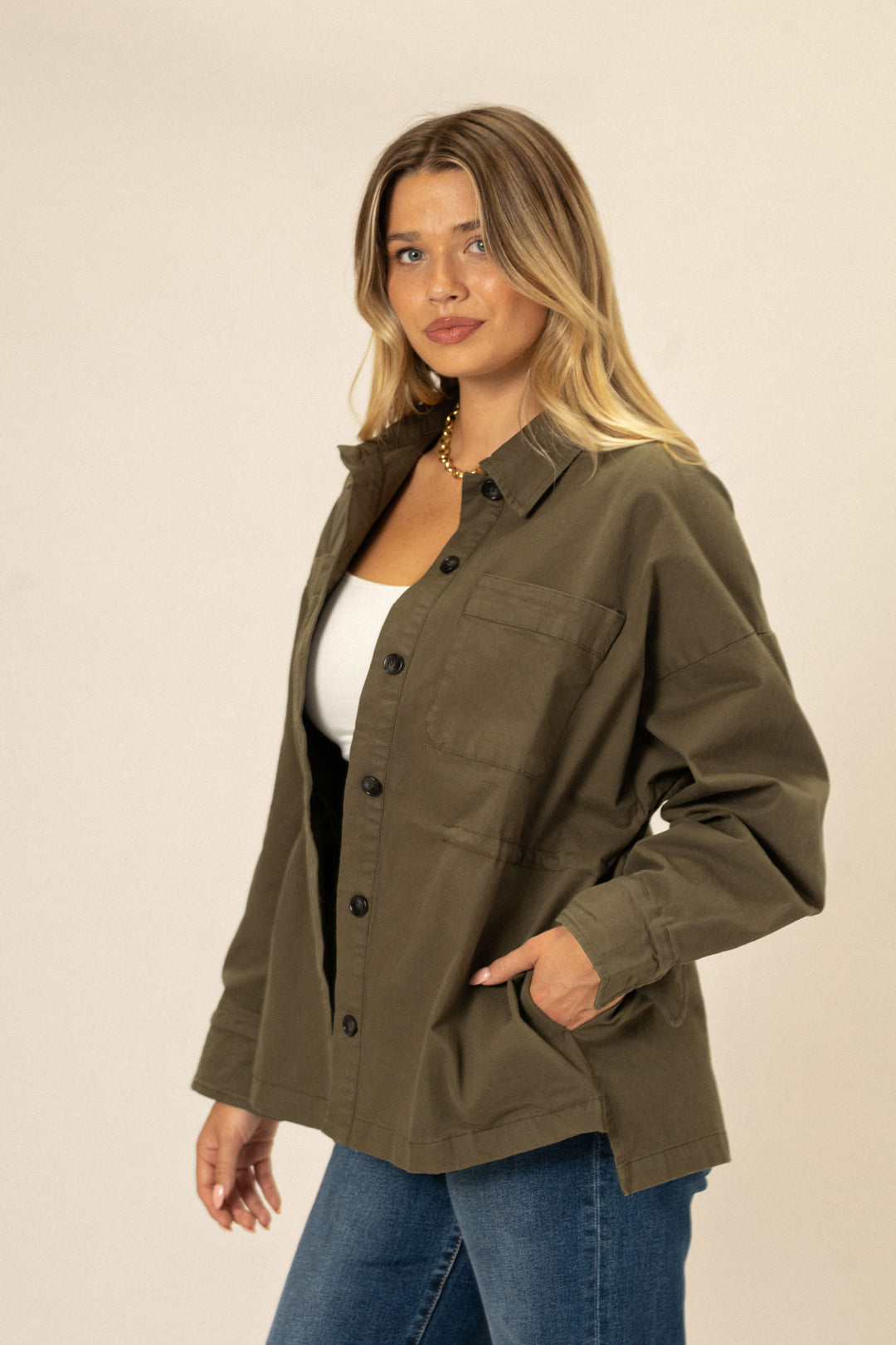 A woman in a khaki green button-up jacket with cinched waist, styled with a white tank top and blue jeans, standing against a neutral background.
