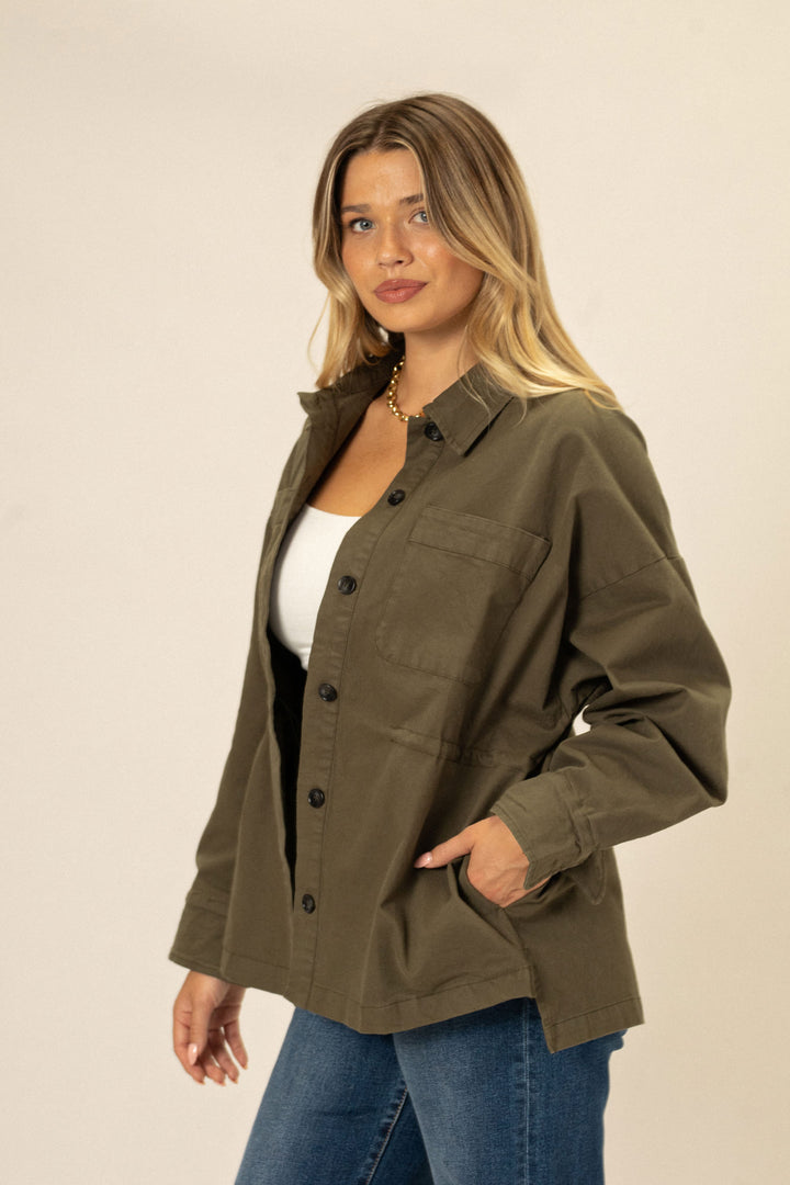 A woman in a khaki green button-up jacket with cinched waist, styled with a white tank top and blue jeans, standing against a neutral background.