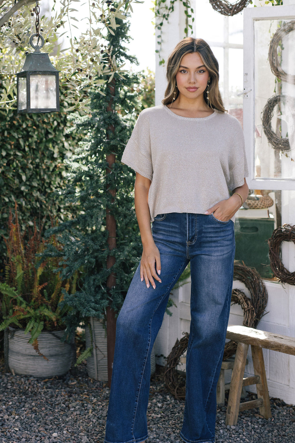 A woman with long, light brown hair wears a metallic beige knit top with a relaxed fit, paired with dark blue denim jeans.