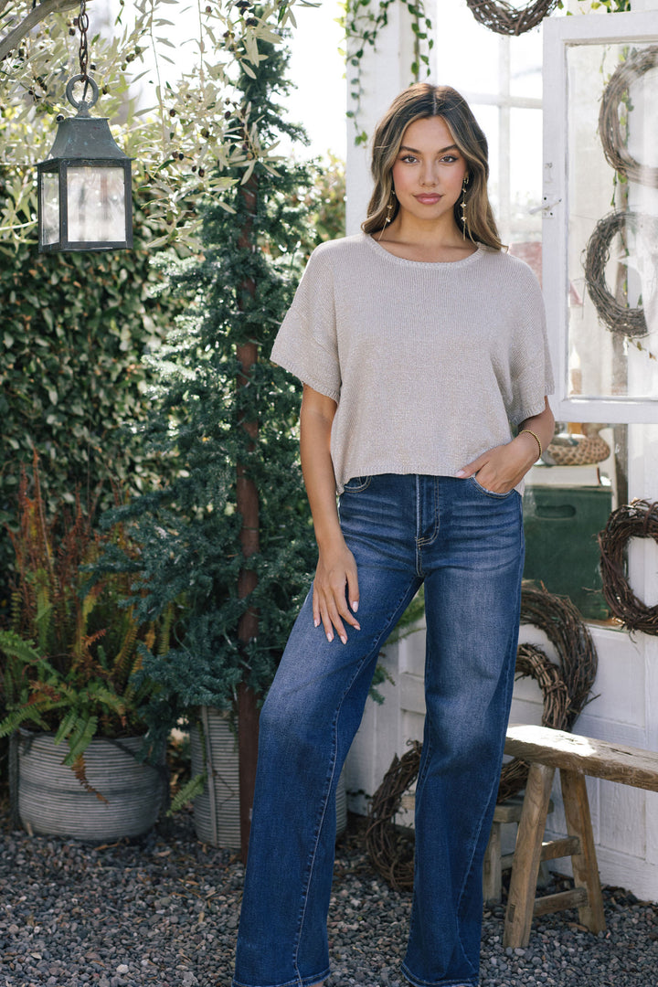 A woman with long, light brown hair wears a metallic beige knit top with a relaxed fit, paired with dark blue denim jeans.