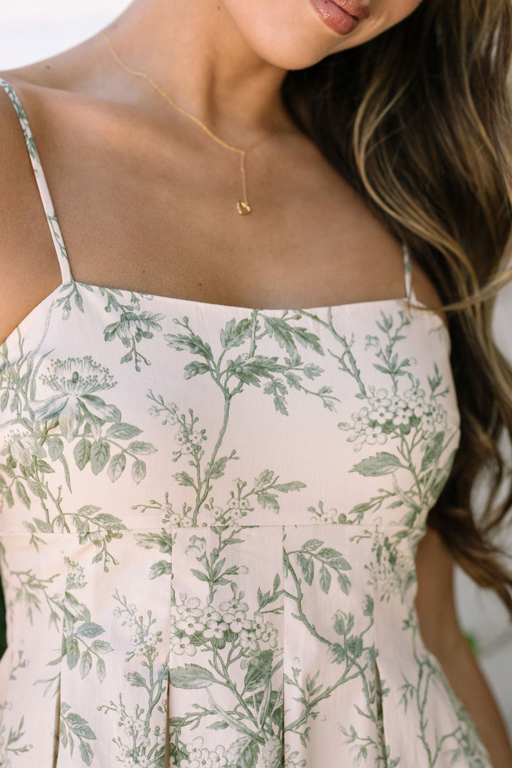 A floral midi dress with a white background and green botanical print, featuring thin shoulder straps, an off-the-shoulder silhouette, and a flowing, knee-length skirt.