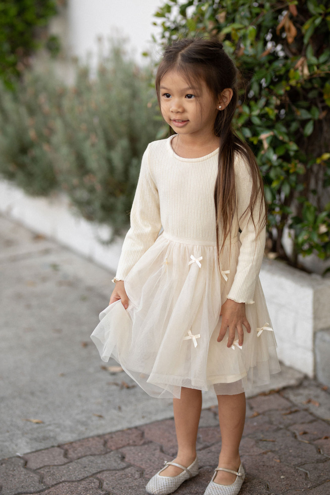 A young girl wearing a white, ribbed knit dress with a tulle overlay and bow details, standing in a natural outdoor setting.