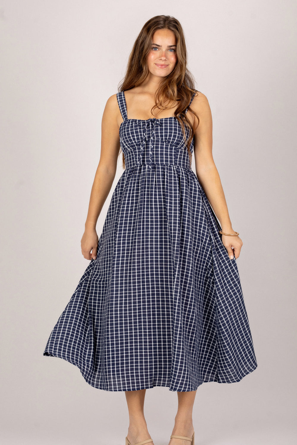 A navy and white plaid midi dress with a fitted bodice, full skirt, and adjustable straps, modeled by a young woman with long wavy brown hair.