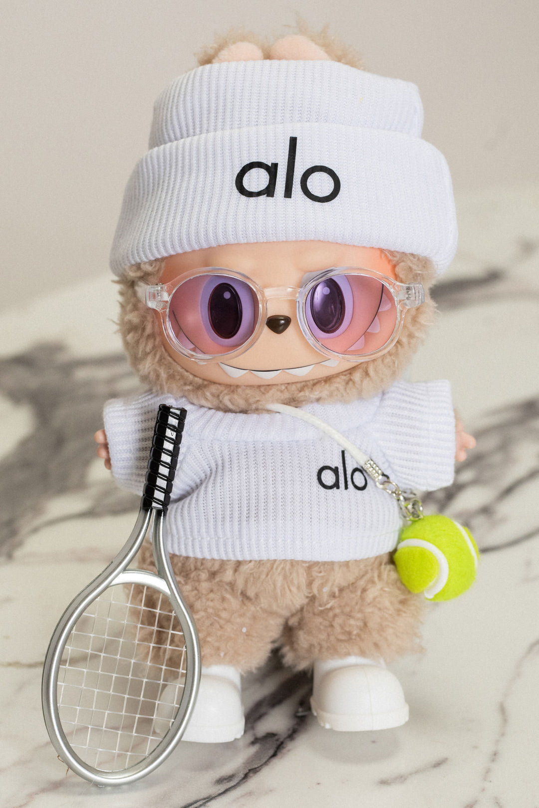 Fuzzy beige plush toy wearing white Alo-branded outfit with sunglasses and holding tennis racket and ball.