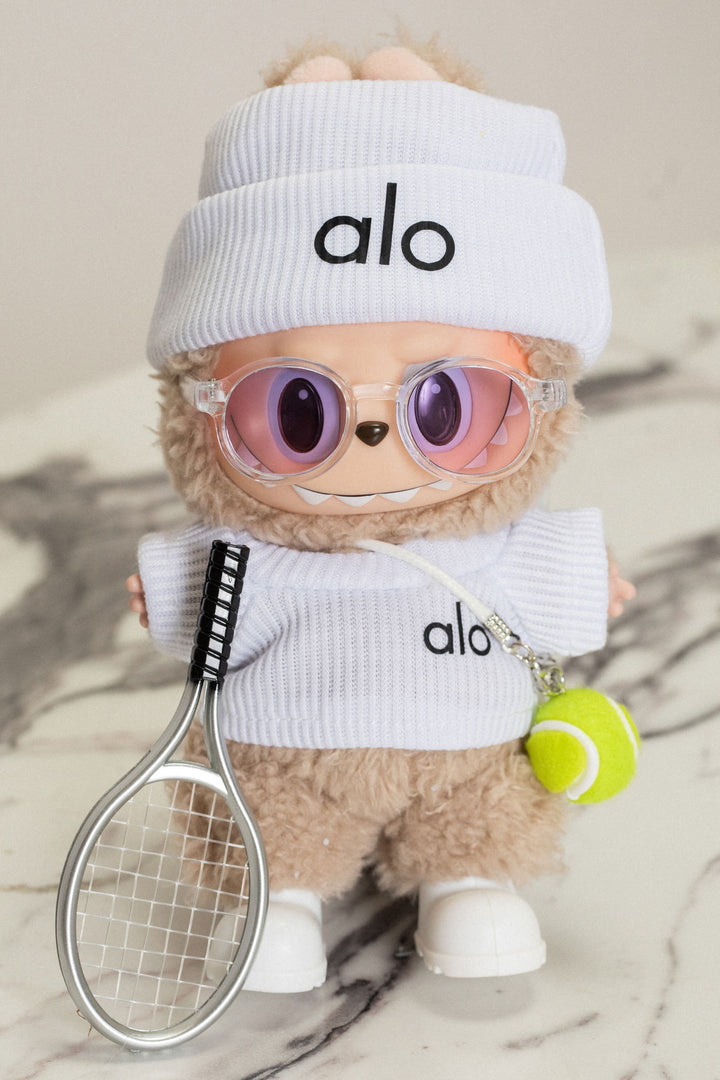 Fuzzy beige plush toy wearing white Alo-branded outfit with sunglasses and holding tennis racket and ball.
