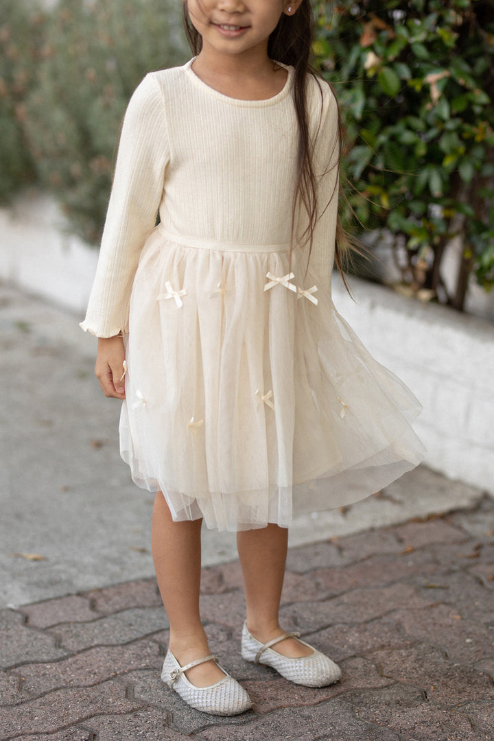 Young girl wearing a white ribbed tulle dress with long bell sleeves and a flared skirt, featuring delicate bows and accents.
