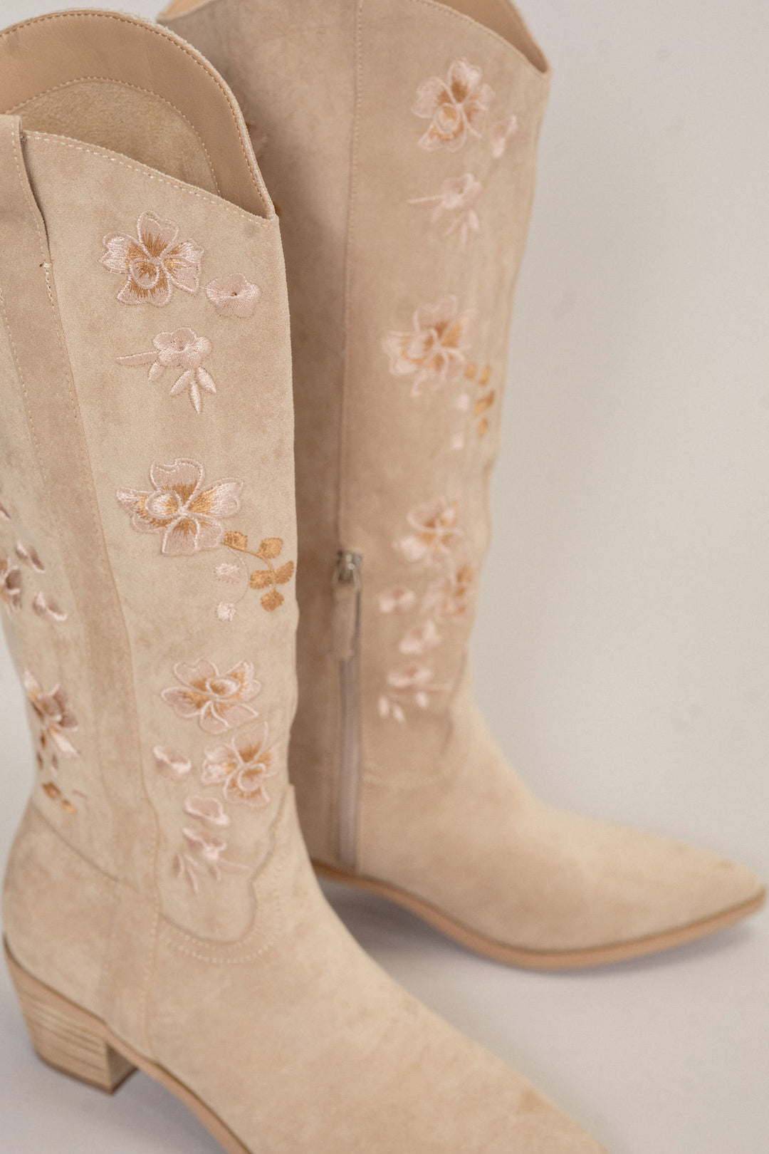 Beige suede cowboy boots with delicate floral embroidered inlay design and zipper closure.