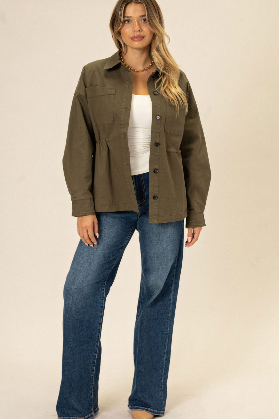 A woman wearing an olive green cinch waist jacket with pockets and button closures, paired with light wash wide leg jeans.