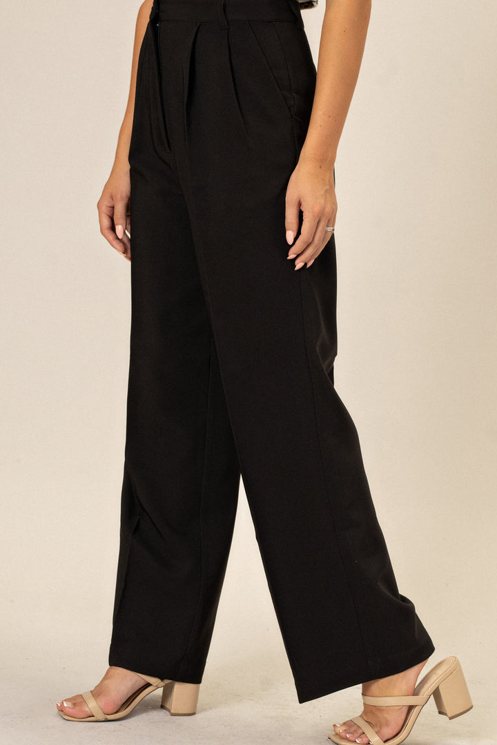 Wide-leg black pants with pleated front detail, paired with beige heeled sandals on a female model.