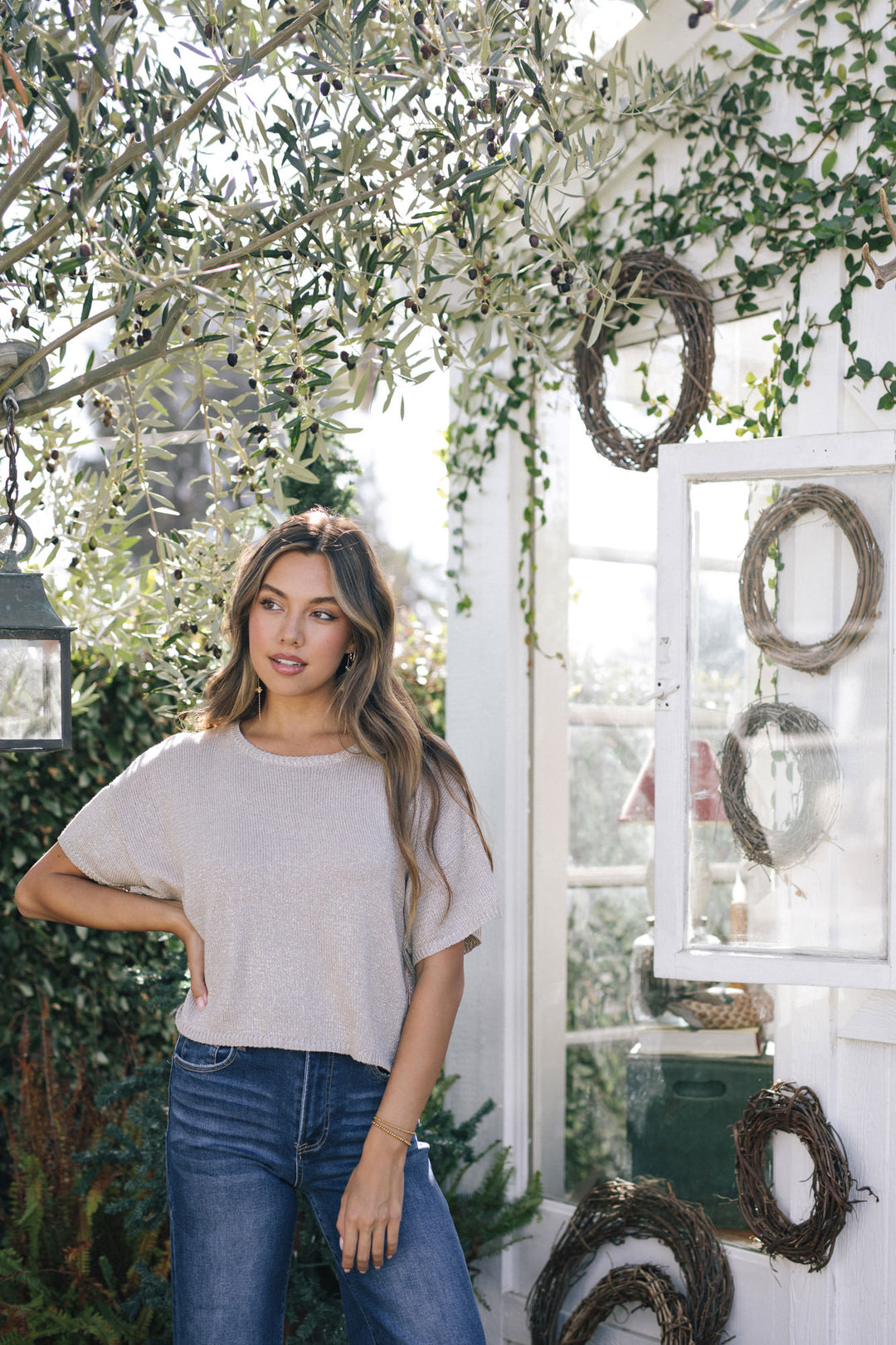 A young woman with long, wavy hair stands in front of an olive tree, wearing a metallic grey knit top and blue jeans. The top features a relaxed, oversized fit and short sleeves.