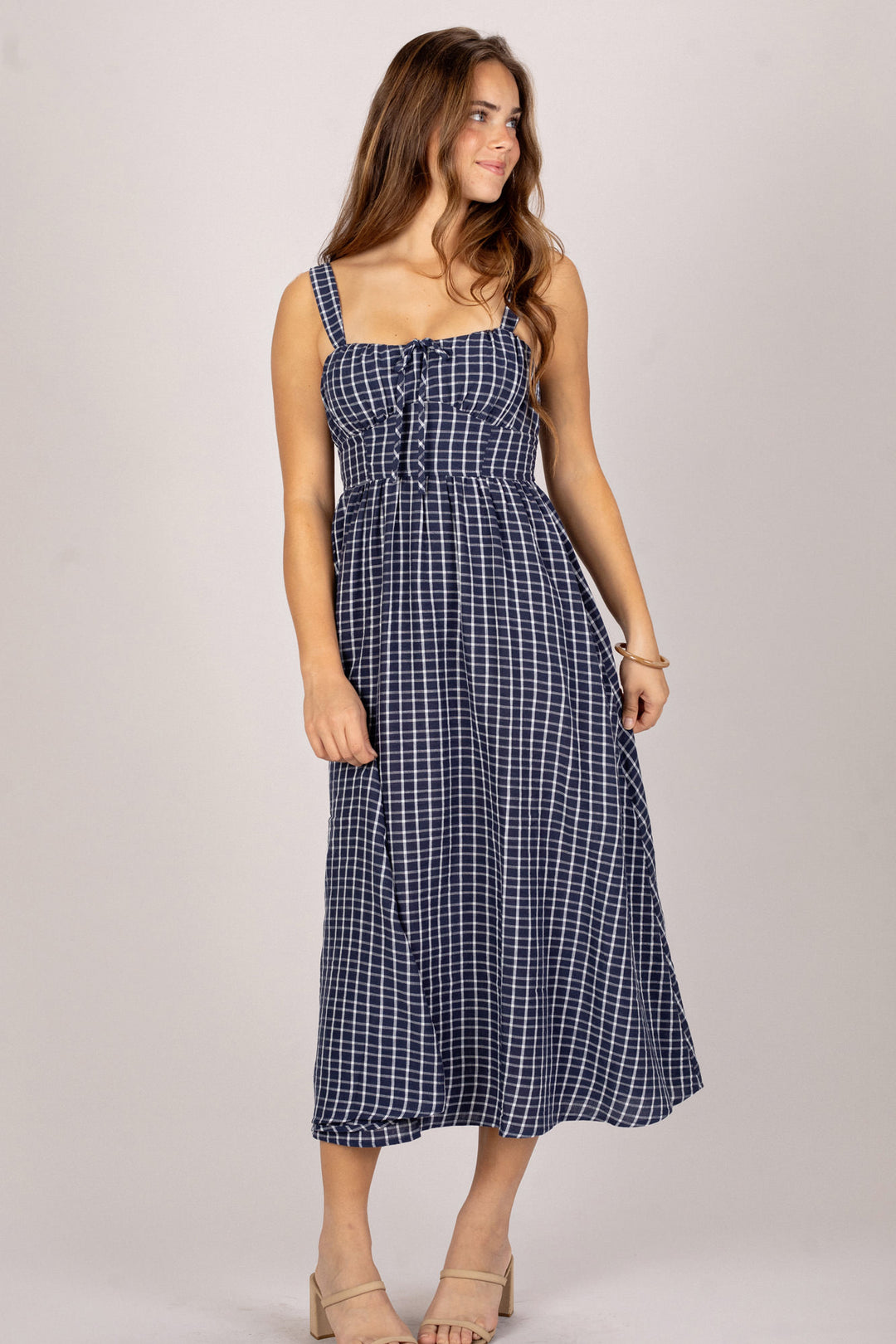 A navy and white plaid midi dress with a ruched bodice, thin straps, and a flared, floor-length skirt, modeled by a smiling woman with wavy brown hair.