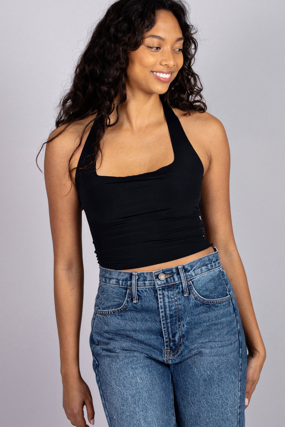 A young woman with curly dark hair wears a black halter-style crop top and high-waisted blue jeans, smiling confidently against a grey background.