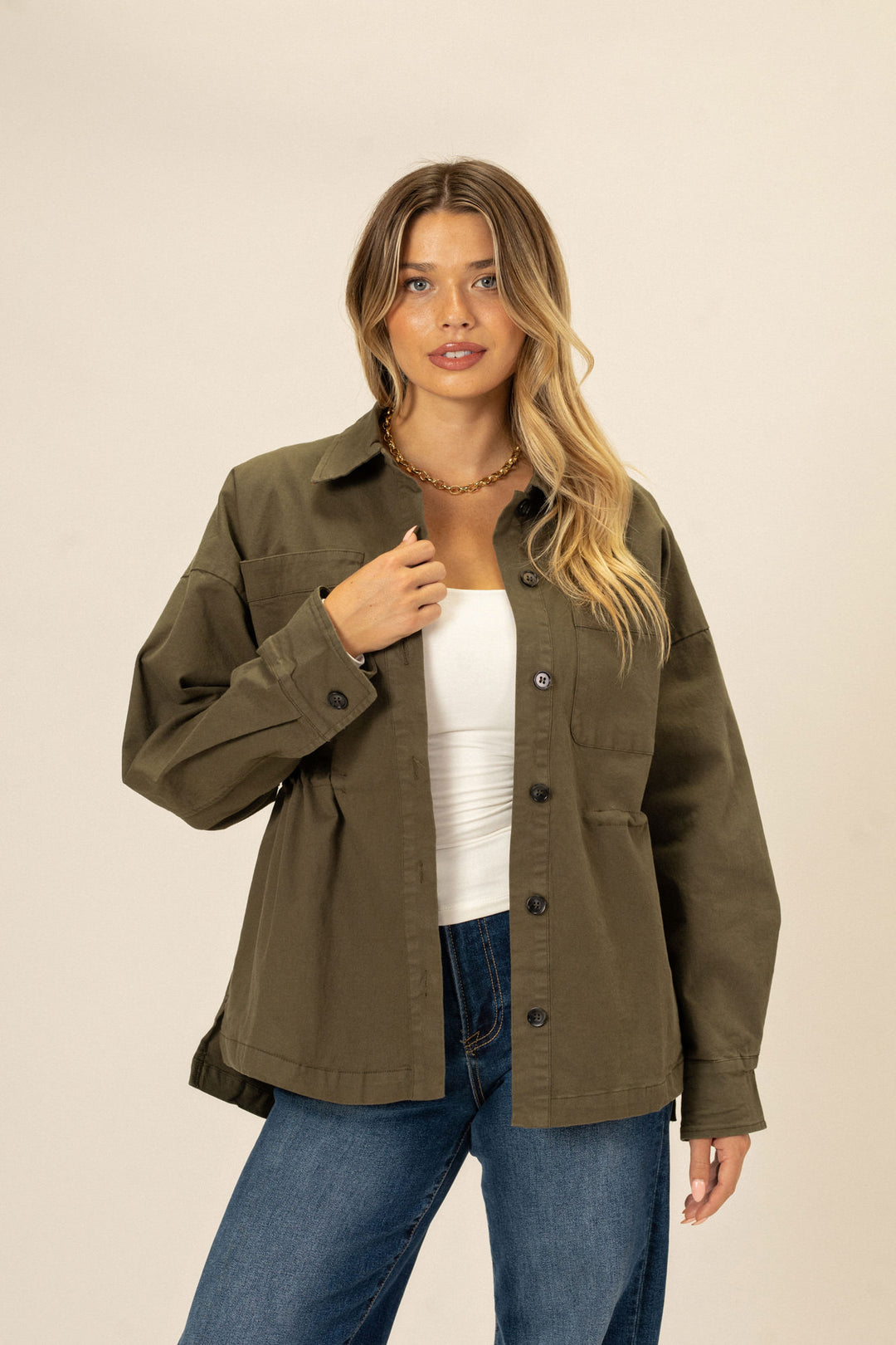 A khaki-colored, button-up jacket with a cinched waist, worn over a white top and paired with blue jeans by a blonde, wavy-haired model.