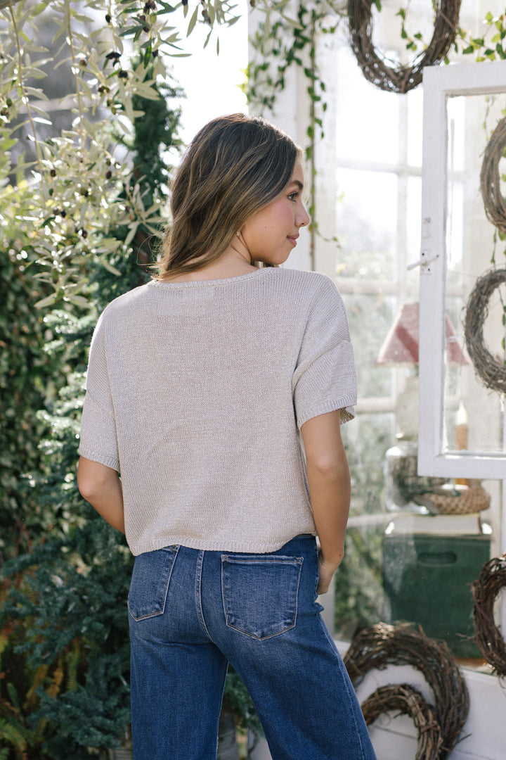 A gray knit short-sleeved top with a relaxed fit and a slightly cropped length, styled with mid-rise blue jeans in a natural setting with lush greenery.
