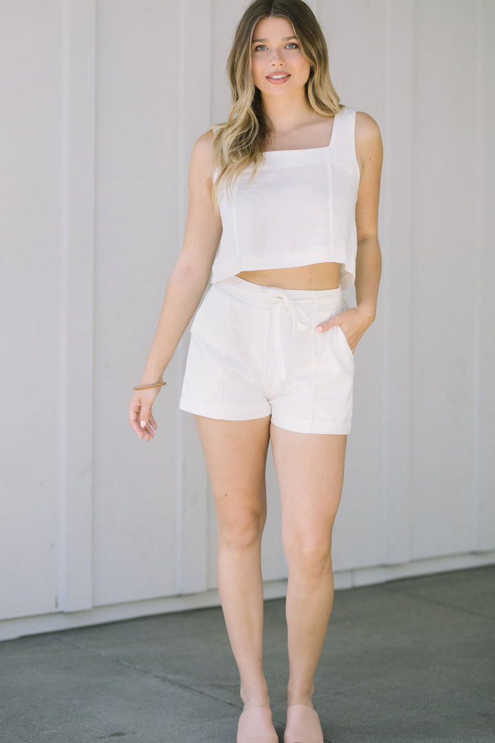 A woman wearing a white square-neck tank top and white shorts, standing in front of a light gray background.