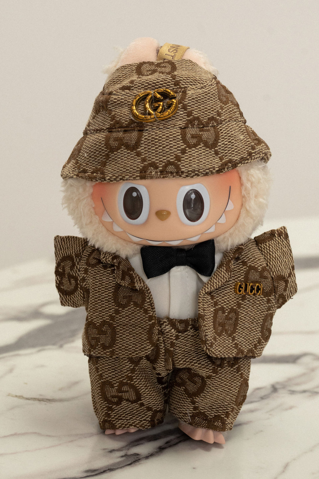 A plush stuffed toy wearing a brown and tan Gucci-branded suit with a bow tie and a Gucci logo cap.