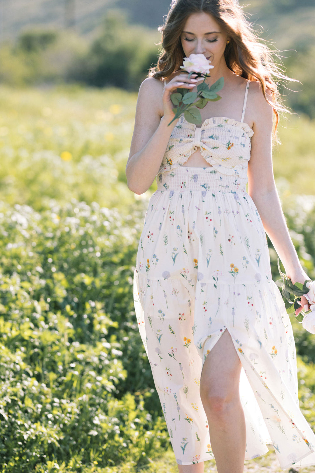 A young woman stands in a field of wildflowers, wearing a white floral print maxi dress with a smocked bodice, flowing skirt, and spaghetti straps.
