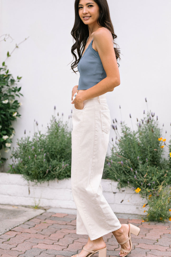 A young woman stands in a garden wearing white barrel jeans, a slate blue ribbed tank top, and beige heels, showcasing the casual yet stylish design of the Drew Barrel Jeans.