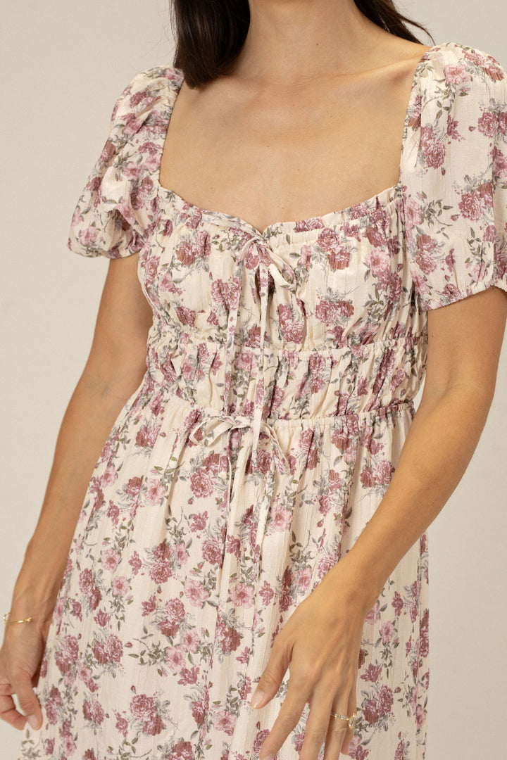 The image shows a woman wearing a floral print dress with puff sleeves and a tie detail at the waist, creating a romantic and feminine look.