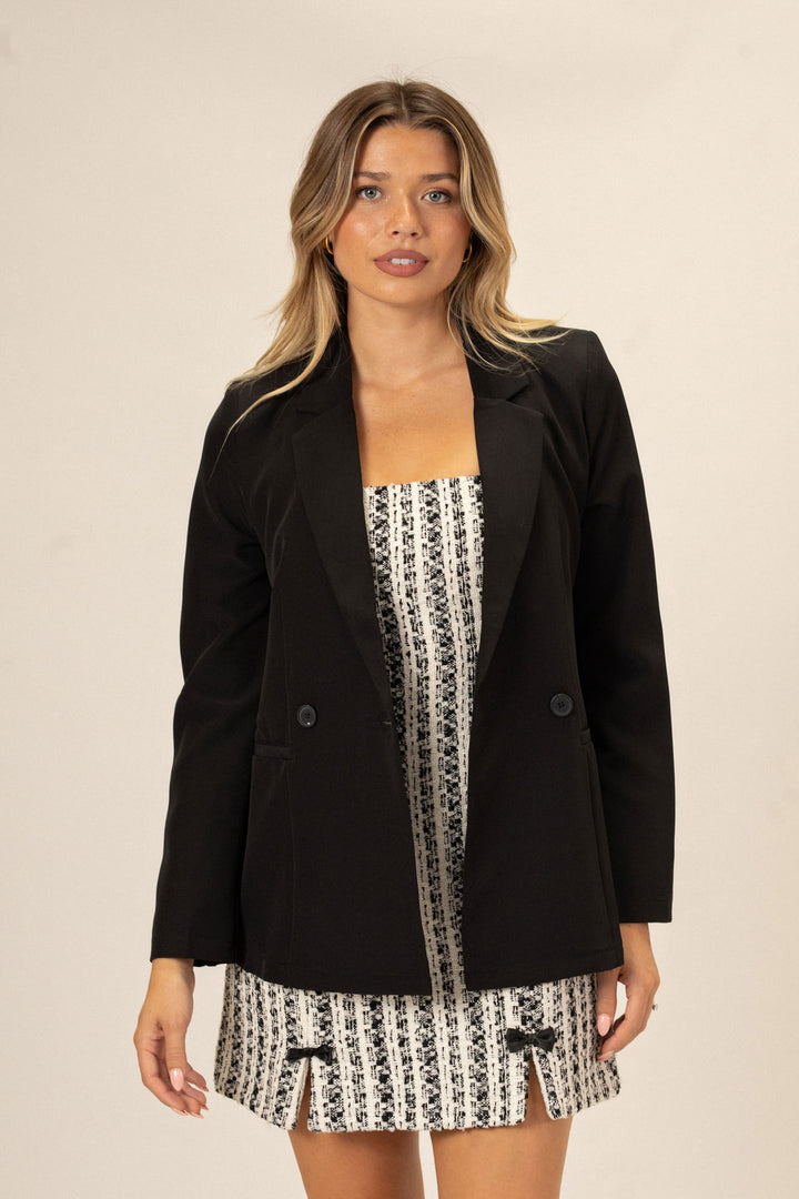 A woman in a black double-breasted blazer over a patterned mini dress, with long blonde wavy hair.