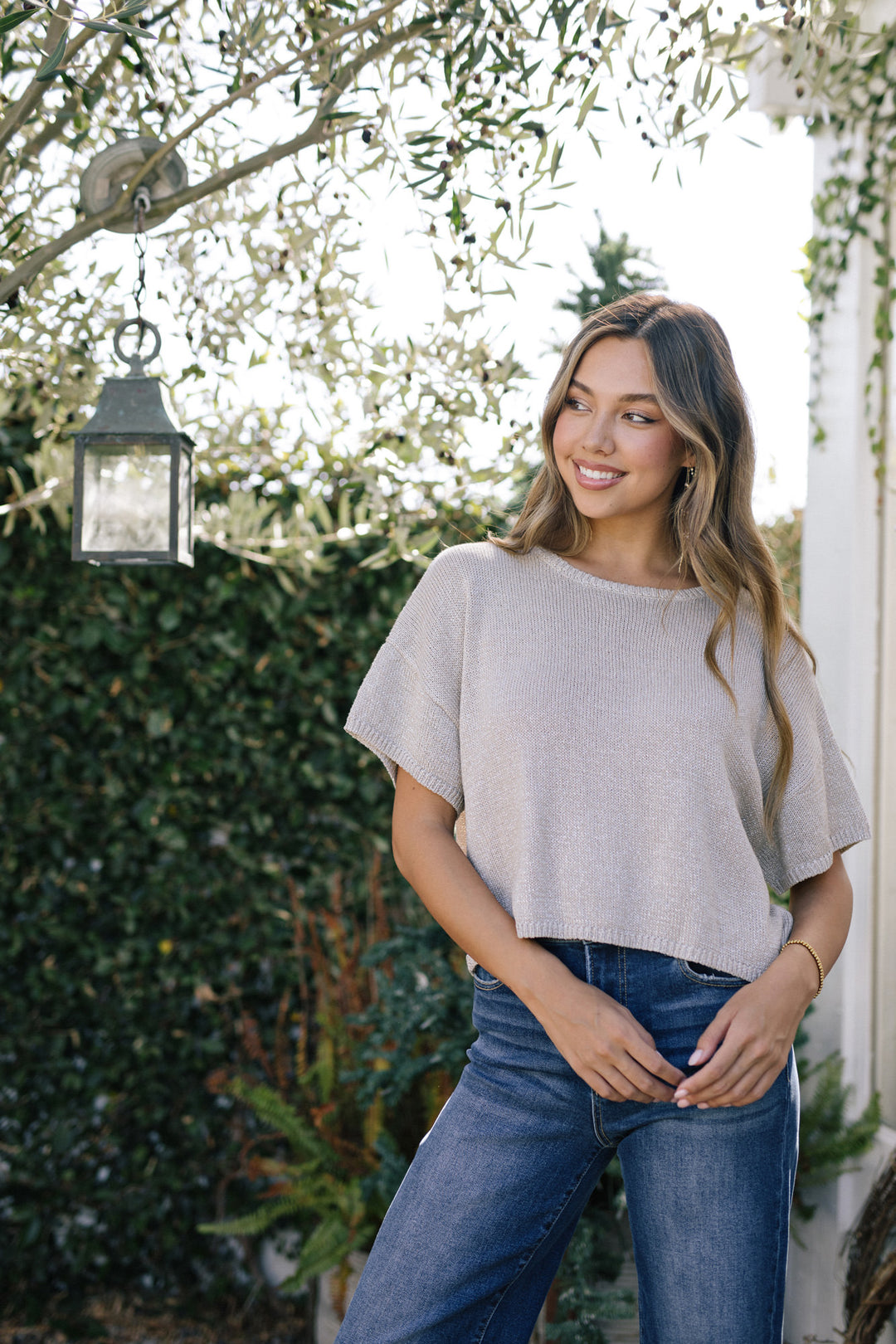 A smiling young woman with long brown hair wearing a beige knit cropped top and blue jeans, standing in front of a lush garden backdrop with a hanging lantern.