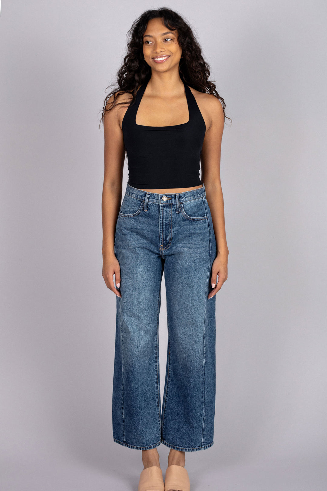 A smiling young woman wearing a black halter crop top and high-waisted light wash denim jeans.