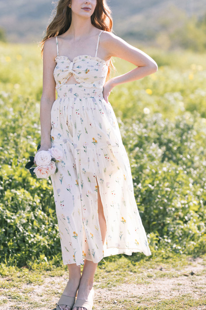 A white floral maxi dress with a smocked bodice, spaghetti straps, and a thigh-high slit, set against a lush, blooming field of yellow flowers.