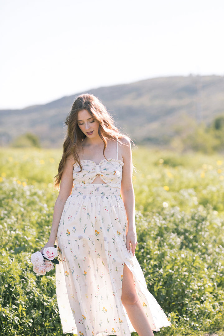 A young woman in a white floral maxi dress with smocked bodice and cutout details, standing in a field of wildflowers against a mountainous backdrop.