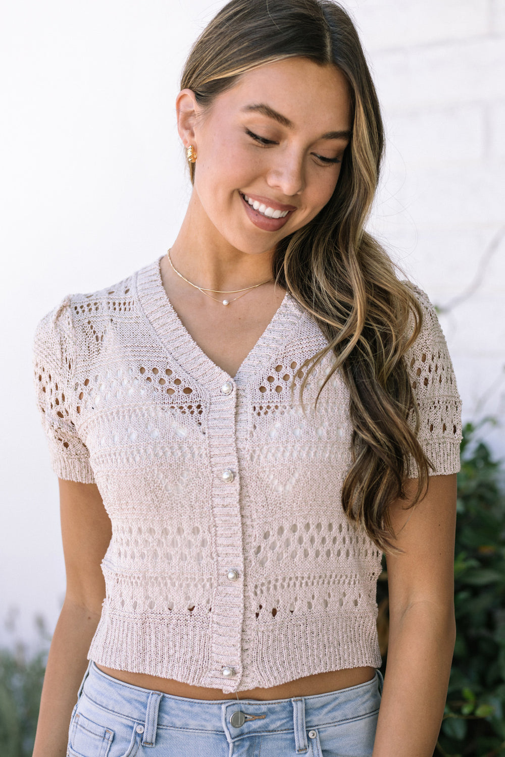 A beige short-sleeved cropped knit cardigan with eyelet and pearl details, modeled by a woman with long wavy hair.