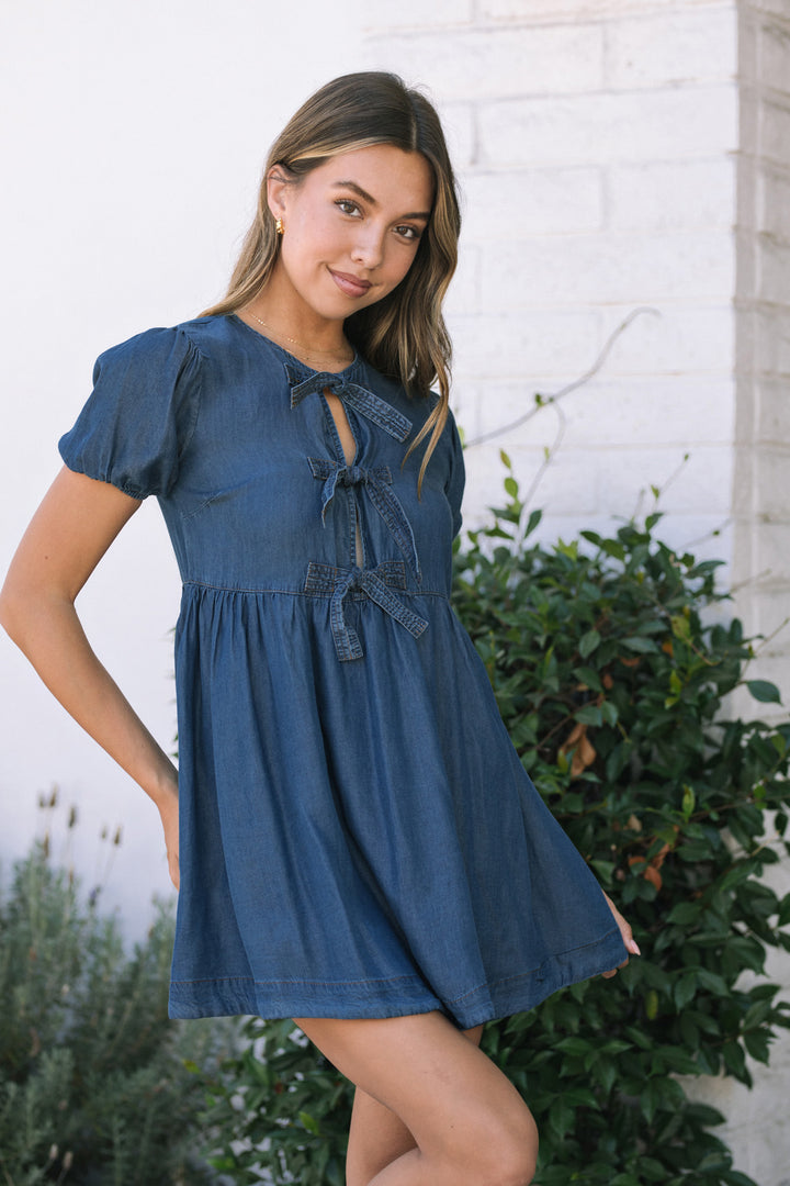Young woman wearing a blue denim dress with puffy sleeves and a bow detail at the neckline, posing in a garden setting.
