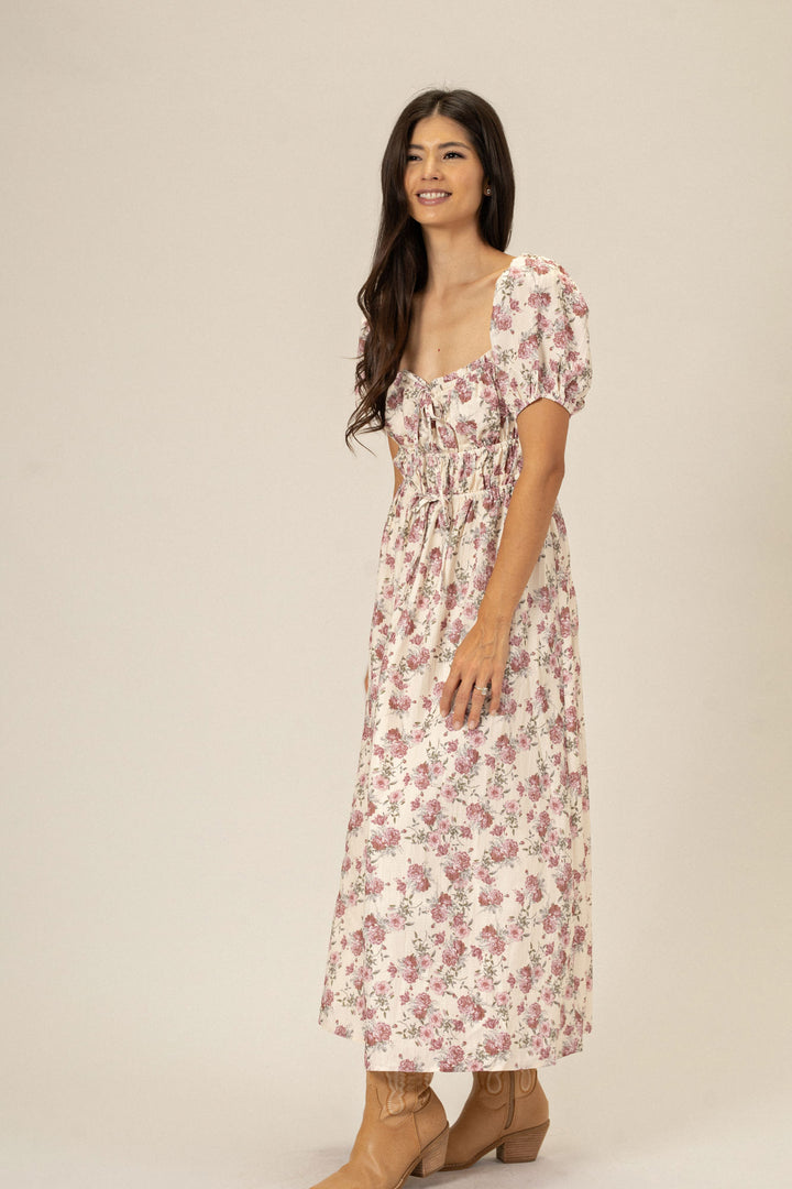 A woman wearing a long, floral-patterned dress with puffed sleeves, standing and smiling against a neutral background.