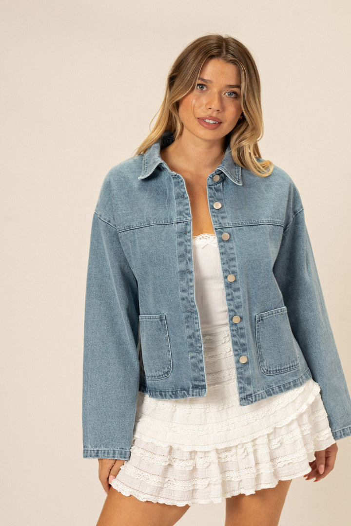 A stylish denim jacket with a buttoned front, in a light blue wash, worn by a model with blonde hair and a white ruffle skirt.