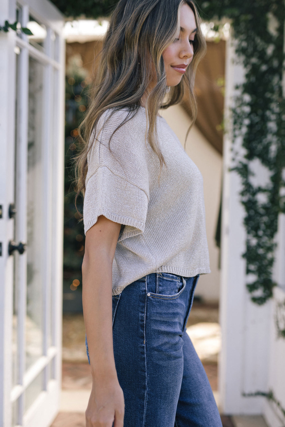A woman wearing a gray metallic knit top with flared sleeves, paired with blue high-waisted jeans, standing in front of a white fence against greenery.