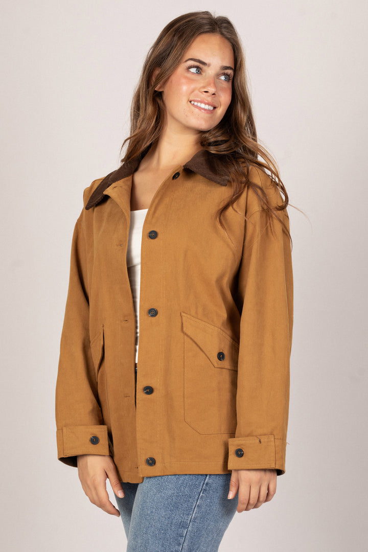 A tan, oversized barn jacket with button closures and pockets, worn by a smiling woman with long, wavy brown hair.