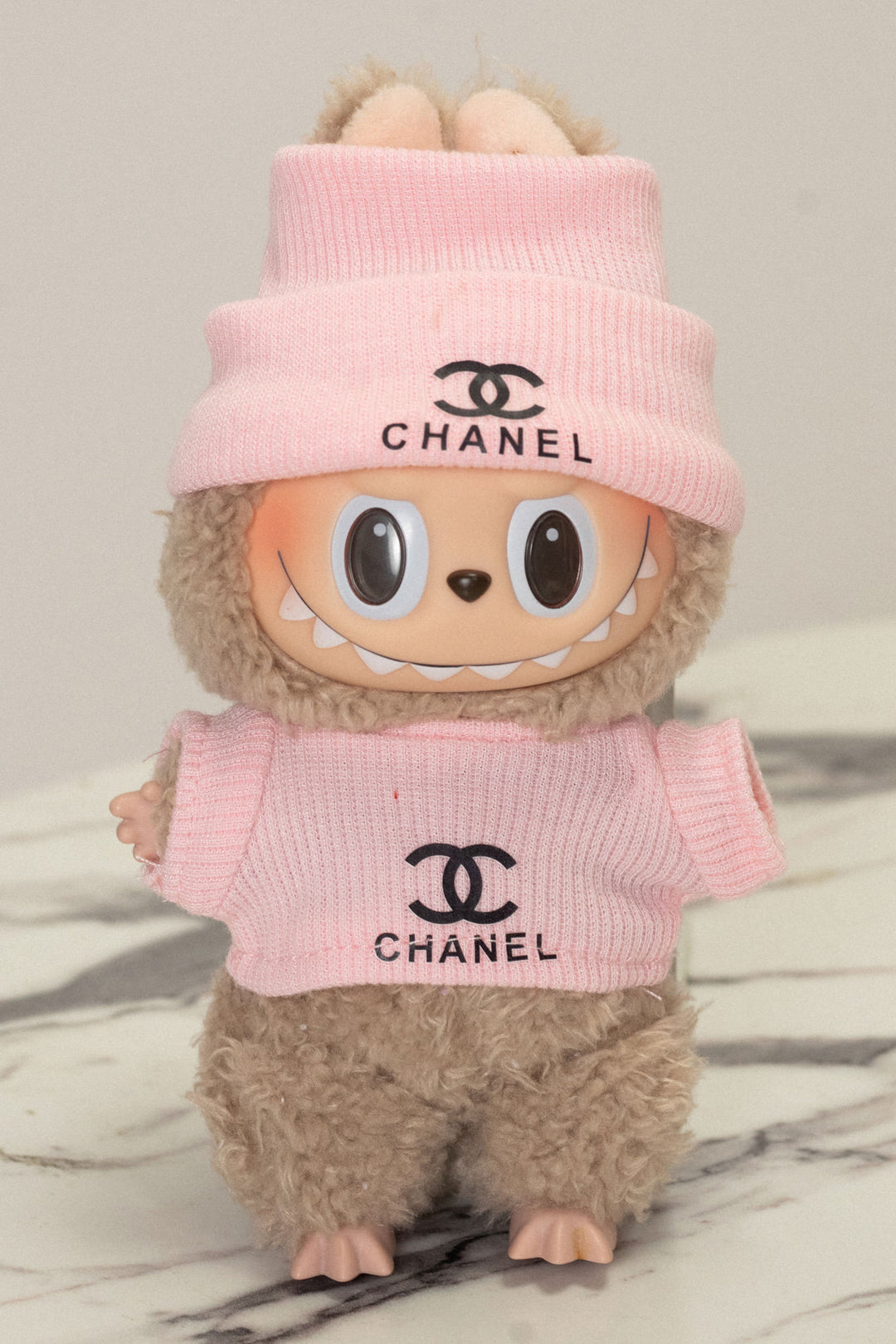A pink, plush Labubu figurine wearing a beanie and top with the Chanel logo.