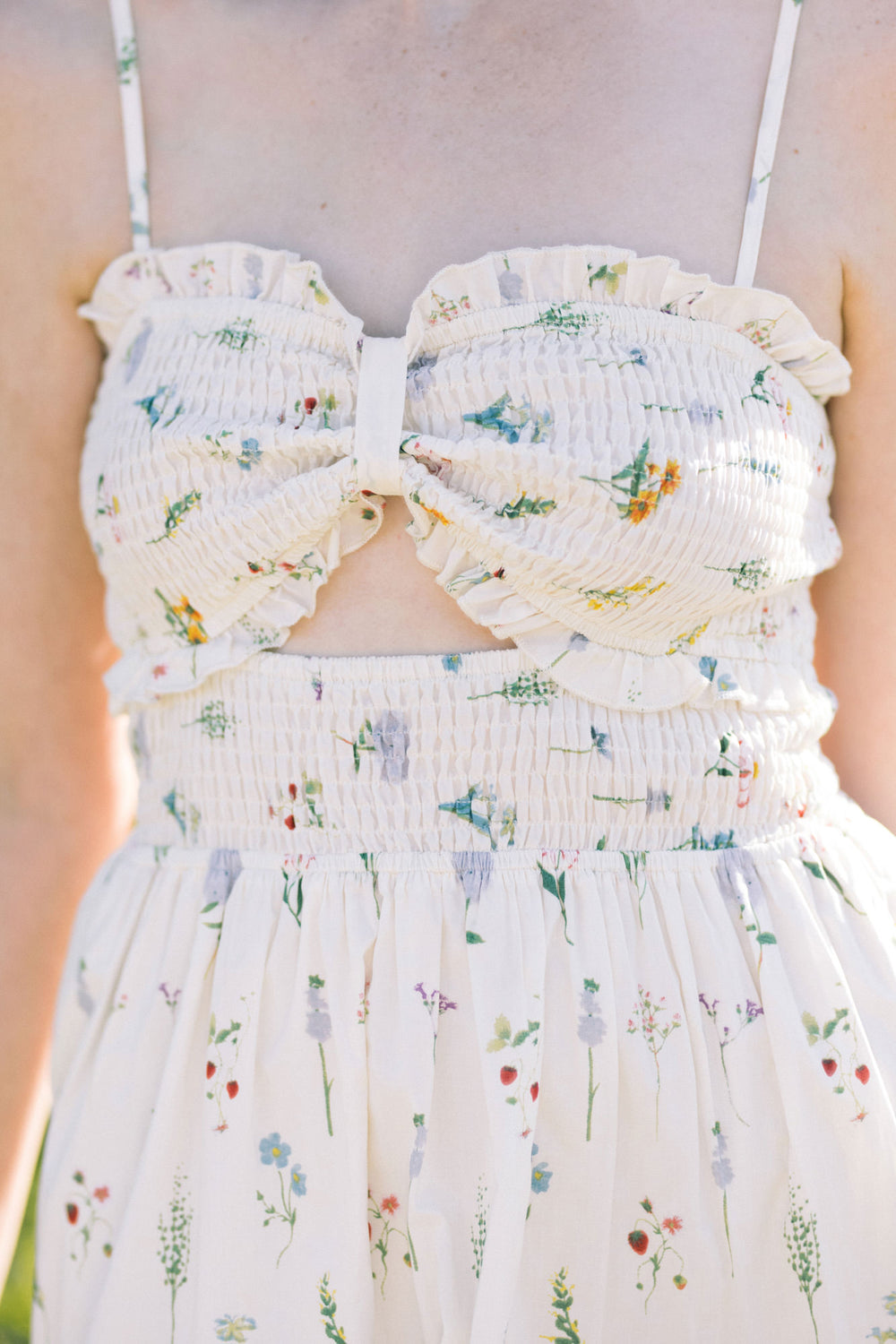 A white sleeveless maxi dress with a smocked bodice and a floral print featuring colorful wildflowers and greenery throughout.