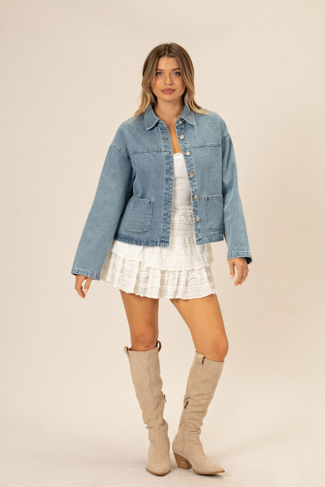 A young woman wearing a light blue denim jacket with button-up closure, paired with a white ruffled mini skirt and beige knee-high boots, standing in a neutral-toned studio setting.
