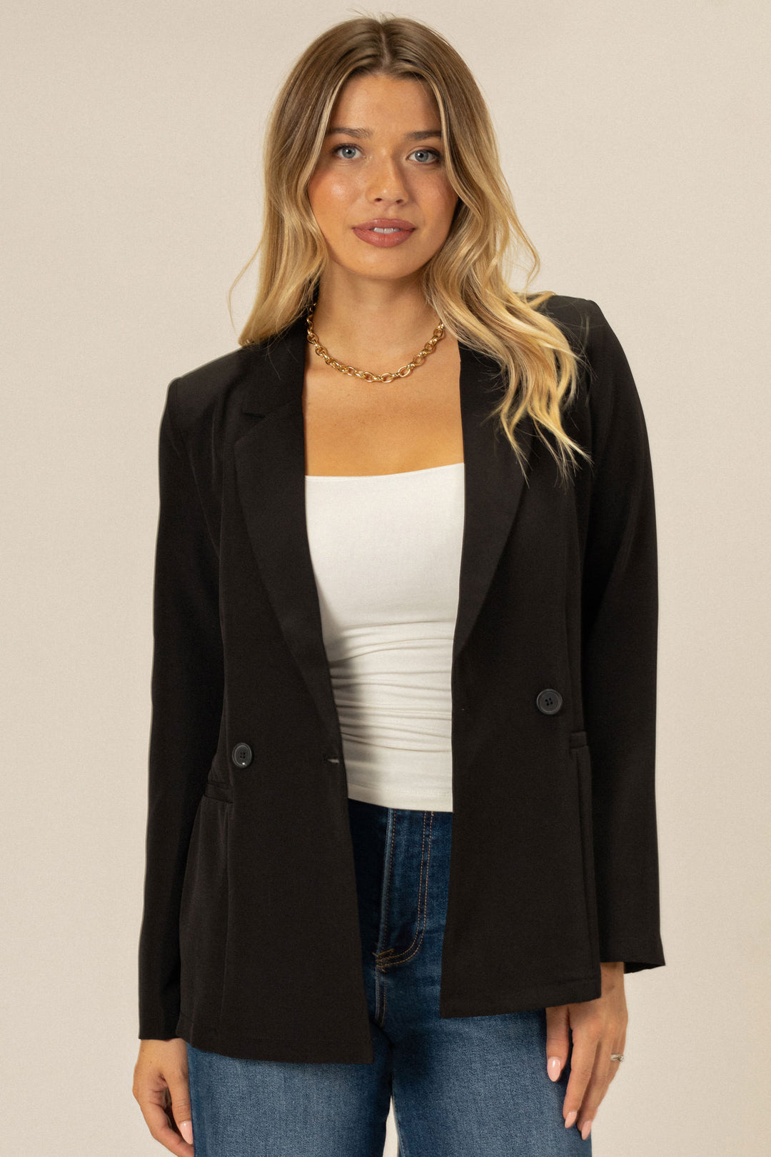 A young woman wearing a black double-breasted blazer with a gold chain necklace, standing against a beige background.
