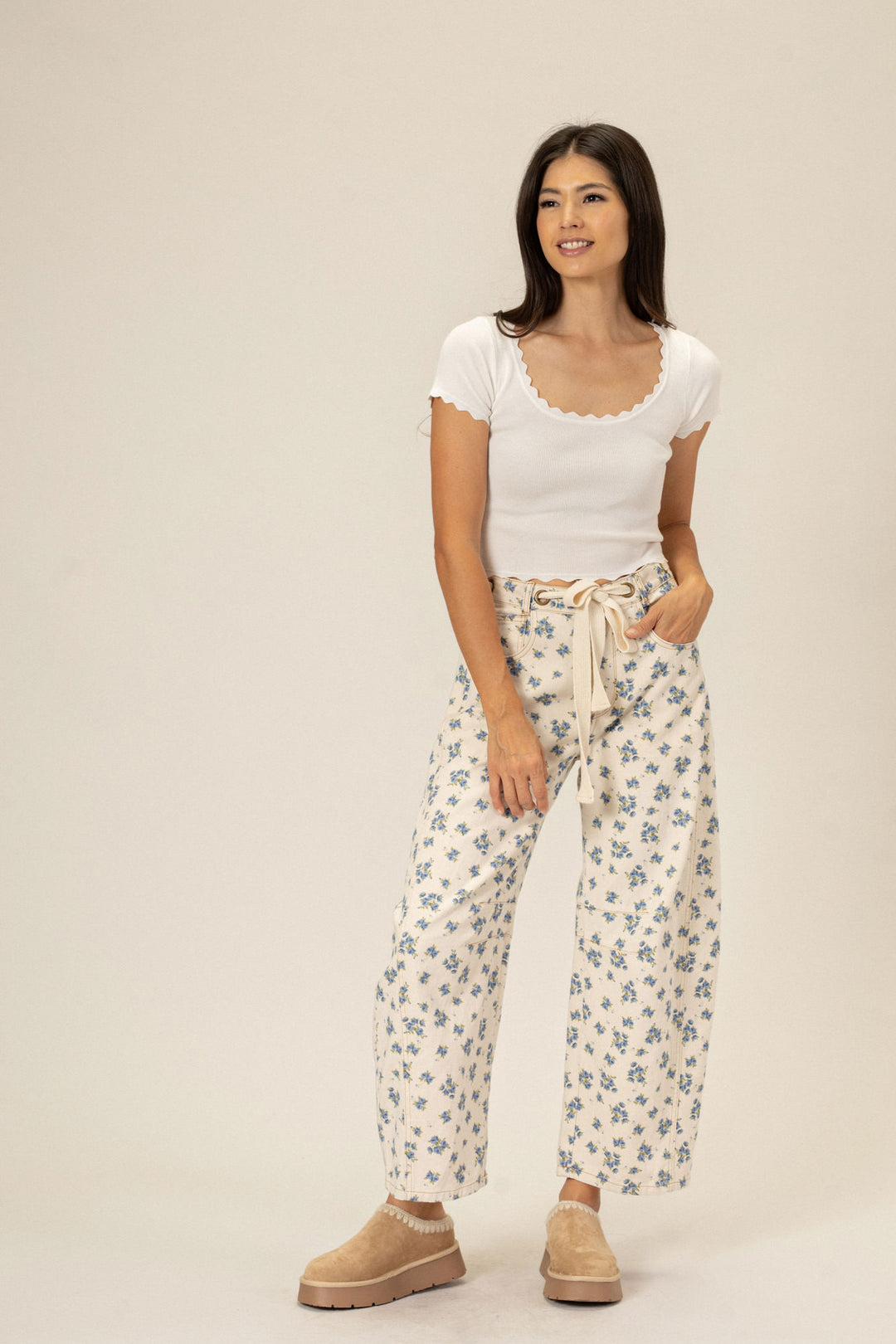 A young woman wearing a white scoop-neck top and beige floral print wide-leg pants, standing against a light background.