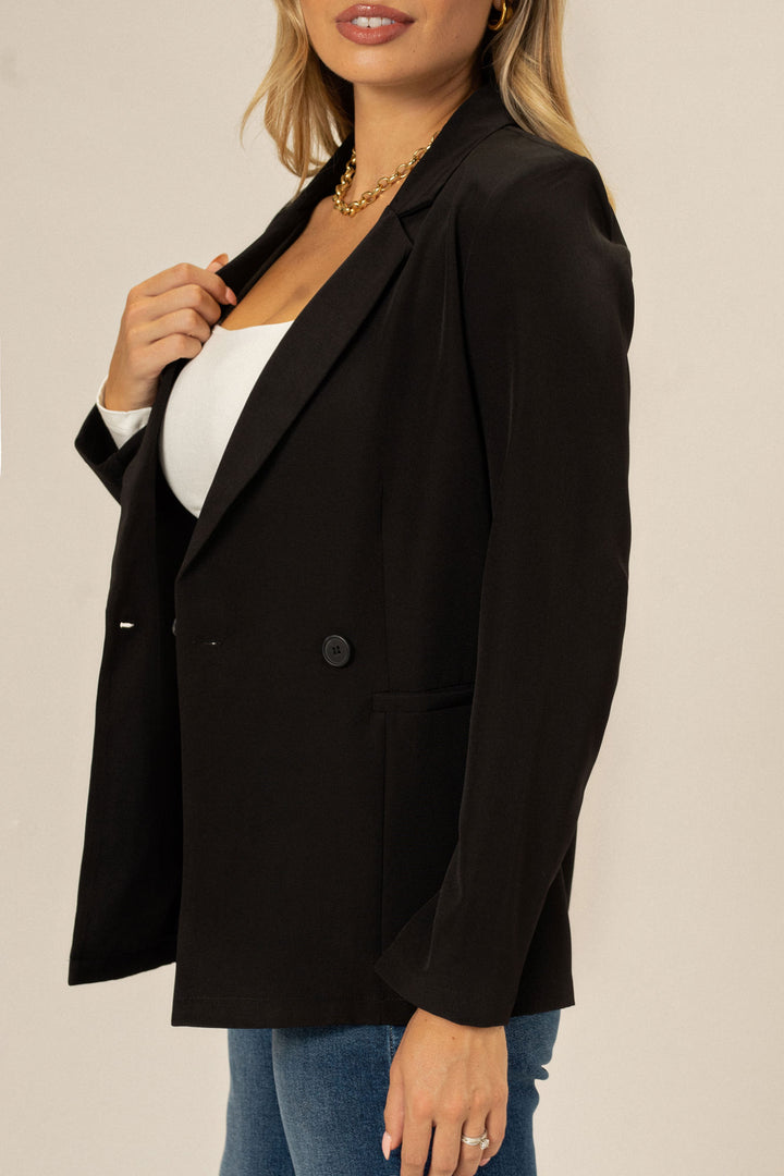 A sleek, black double-breasted blazer with structured shoulders and a tailored fit, styled with a white top and gold chain necklace.