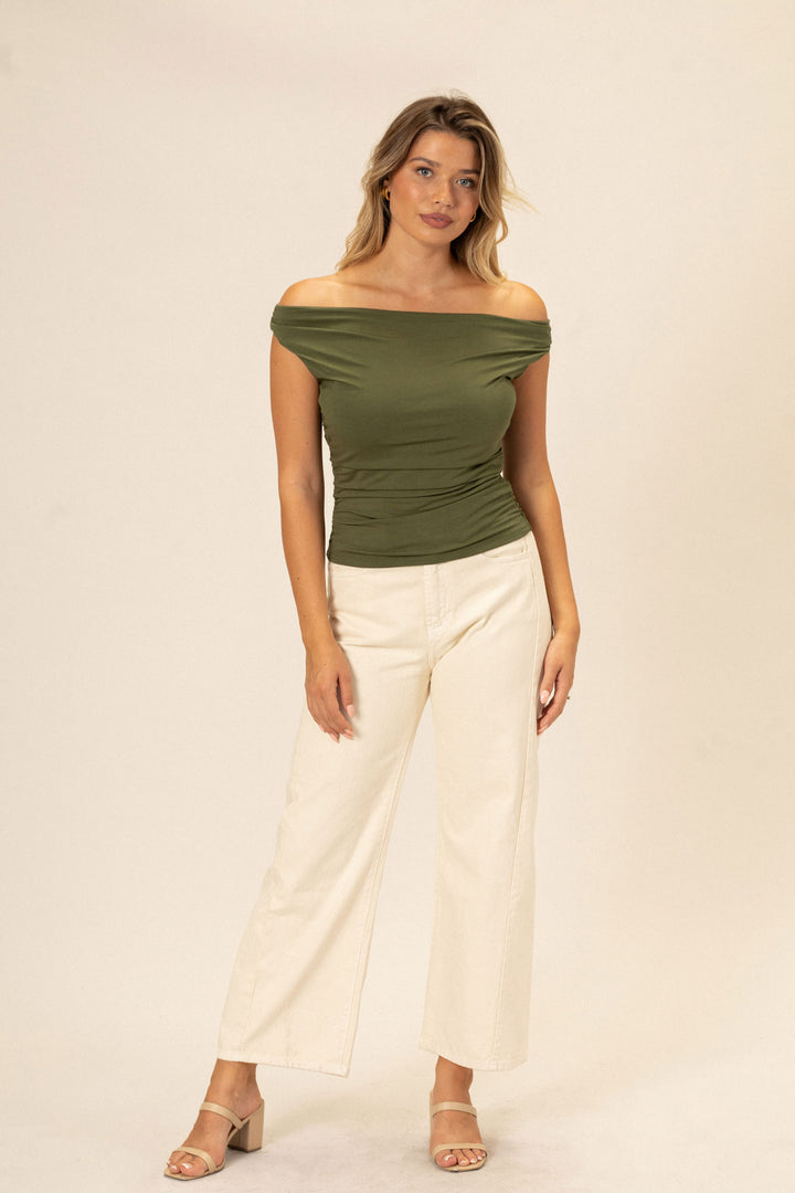A model in a fitted olive green off-the-shoulder top, paired with high-waisted white pants and tan sandals, showcasing the chic, versatile design of the Chriselle Top.