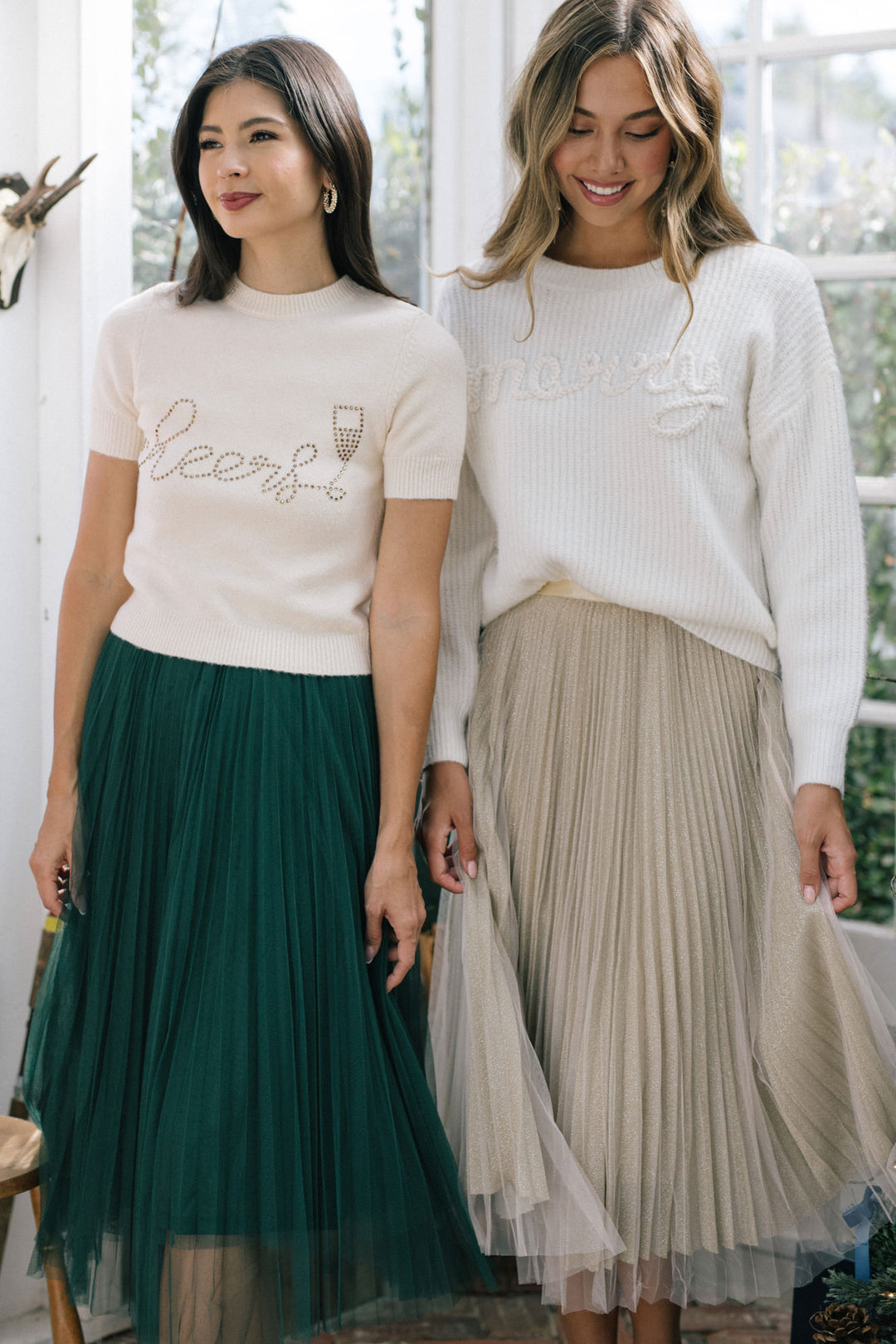 Two women wearing white sweaters with text and patterned skirts, posing in a bright, airy setting.